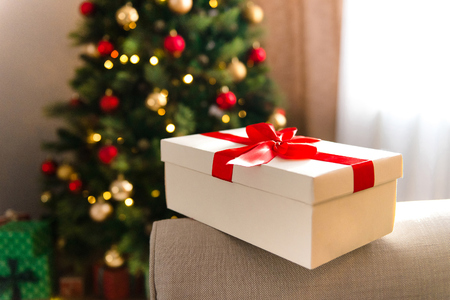 Close view on a cute Christmas present in a white packing with a bright red ribbon, new year magic atmosphere and a Christmas tree in the corner of the room, copy spaceの写真素材