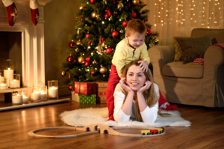 Cute little boy is climbing his lying mom's back, new year traditionally decorated room. Merry Christmas and Happy New Year. Beautiful young mother with sonの写真素材