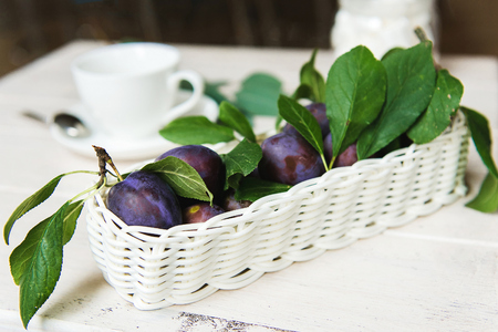 White wicker filled with fresh plums and green leaves. Ripe purple plums with leaves in metal decorative basket over white wooden backgroundの写真素材