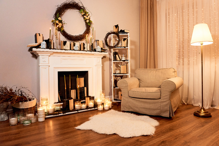 Autumn interior in a cozy house. Fireplace with candles, cozy armchair, autumn wreath on the wall. Night windows at the backgroundの写真素材