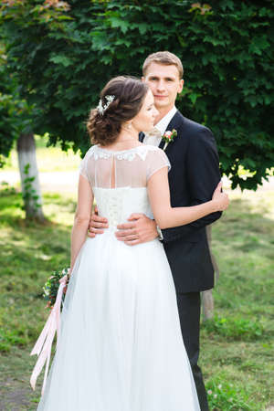 Young couple just married. Wedding couple hugging gently in a green garden, the bride standin back to the shotの写真素材