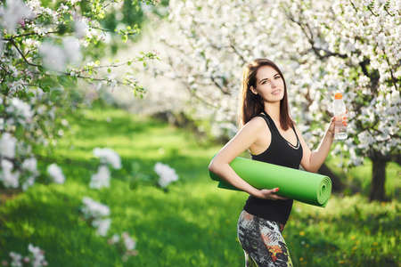 Young smiling positive woman going workout in garden holding water bottle and fitness mat. Active lifestyle candid fitness concept.の写真素材