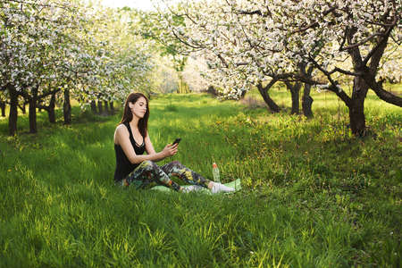 Sporty attractive young woman sitting on a green lawn in the blooming garden, holding the phone in one hand, listening to music on wireless headphones, smiling. Relaxing calmness concept.の写真素材