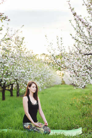 Young caucasian yogi woman with meditating seated during morning exersize on the green lawn in blooming garden, practicing breathing yoga in Virasana, outdoors meditation reduces mental fatigueの写真素材