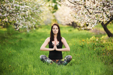 Newbie smiling yogi girl with closed eyes wearing sportswear, sitting on the green lawn in spring blooming garden, practicing yoga in half Lotus pose with namaste, full length.の写真素材