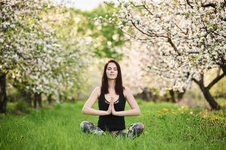 Harmony concept. Smiling yogi girl with closed eyes wearing sportswear, sitting on the green lawn in spring blooming garden, practicing yoga in half Lotus pose with namaste, full length.の写真素材