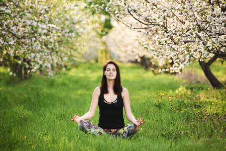 Yogi girl with closed eyes sitting on the green lawn in spring blooming garden doing Ardha Padmasana exercise, full length.の写真素材