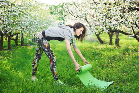 Happy young woman getting ready for workout and unrolling her exercise mat outdoor, blooming garden background.の写真素材