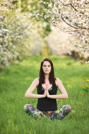 Yogi girl with closed eyes sitting on the green lawn in spring blooming garden, practicing yoga, doing exercise with namaste, full length. Vertical photographyの写真素材