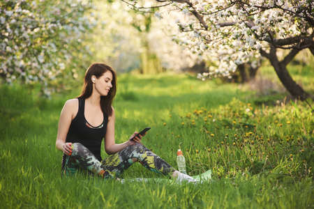 Young woman sitting on a green lawn in the blooming garden, holding the phone in one hand, listening to music on wireless headphones, smiling. Relaxing calmness concept.の写真素材