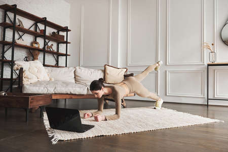 Young attractive woman working out at home interior, doing easy plank exercise keeping fit with abs at home, using online fitness daily training program for beginners on laptop, side view.の写真素材