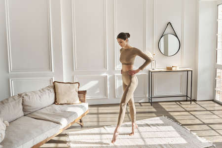 Full length portrait of tall, slender young woman doing morning exercises in living room for flexability, training legs. Fitness at home, lifestyle and personal care concept, working out, sportswearの写真素材