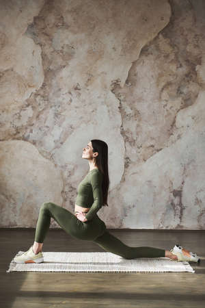 Young attractive woman with long dark hair wearing olive-colored sportswear, working out in yoga studio near grunge wall background. Half splits exercise, practicing yoga. Healthy lifestyle conceptの写真素材