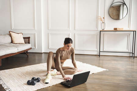 Young brunette woman in beige sportswear is sitting on the floor with dumbbells is using a laptop at home in the living room. Sport and recreation concept. Watching Video Tutorial On Laptopの写真素材