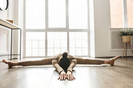 Young woman working out at home, doing Wide-Angle Seated Forward Bend pose, doing split exercise at...