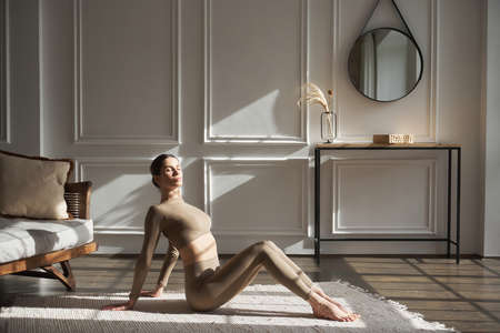 Young slim elegant woman in beige sportswear is lying on a natural rug on the floor, resting and relaxing after training, full length, side view, white yoga studio. Wellness, harmony concept.の写真素材