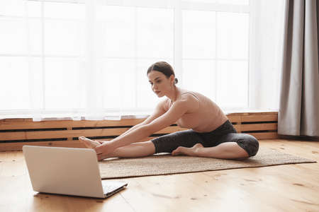 Young woman practicing yoga stretching, watching online video tutorials by professional fitness instructor, exercising from home because of social distancing. Concept of online learning, full lengthの写真素材