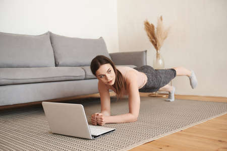 Attractive female doing planking bodyweight exercise and watching online tutorials on laptop, training in the living room, copy space. Online training on laptop computers. Concept of online learning.の写真素材