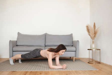 Sporty young woman doing morning exercise alone in living room stands in plank leaning on her elbow. Workout at home for a healthy body, lifestyle concept. Copy space, full length, side viewの写真素材