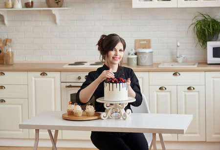 Young professional confectioner happy with her work, carefully corrects the berries on the cake in the kitchen, looking at camera. Lifestyle cooking concept. Copy Space.の写真素材
