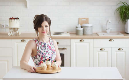 Young woman sitting at the table, holding a tray with a delicious cupcake with cream and berries in a white kitchen. Weight Loss Concept. Copy spaceの写真素材