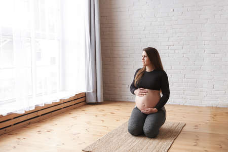 Young smiling pregnant woman sits on her knees a natural rug, doing hip stretching exercises, holds tummy, looks in the big window, rest after yoga exercise. Horizontal, Full length, copy space.の写真素材