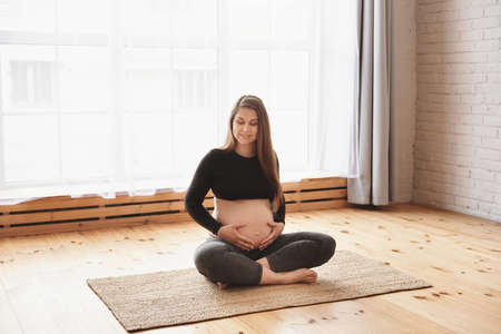 Portrait of young smiling pregnant woman rest after working out gently hugs her tummy indoors. Pregnant fitness person smiling while practicing yoga at home. Prenatal relaxation with closed eyesの写真素材