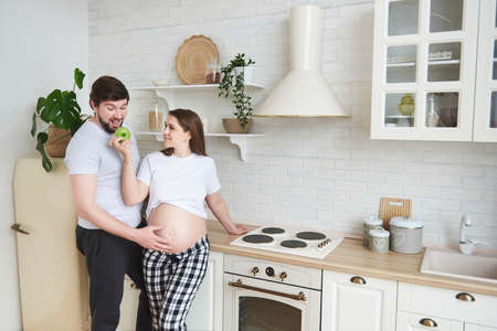 Cute and funny future parents in home outfit embrace standing in the kitchen. Pregnant wife gives husband a green apple. Healthy Lifestyle, parenthood, maternity, expectation of a child, copy space.の写真素材