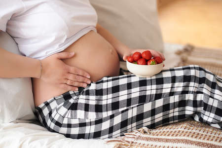 Pregnancy, healthy food and people concept. Beautiful tummy of a pregnant woman in home outfit, lying on a bed with a strawberries plate. Close up, copy space, alergia, diet and healthy food conceptの写真素材