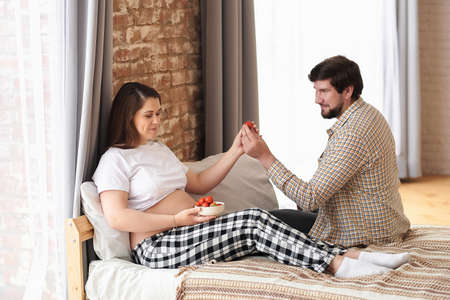 Romantic husband on the bed, giving strawberries to his pregnant wife. Healthcare and healthy food. Happy family pregnancy, wellbeing in family, affectionate couple portrait, and expectation concept.の写真素材