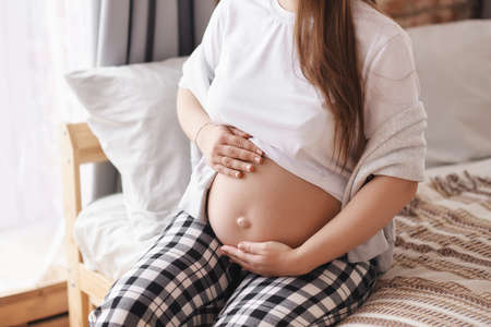 Close-up pregnant woman in home outfit: a white T-shirt and checkered pants sits on the bed, hugs her tummy. Prenatal, Baby expecting concept, studio, copy space.の写真素材