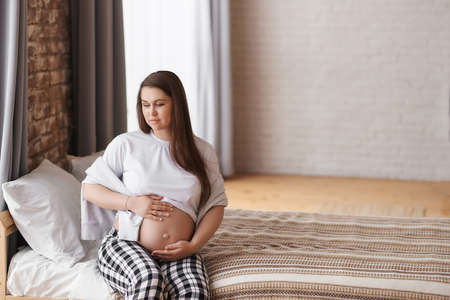 Calm pregnant woman looking down in home outfit: a white T-shirt and checkered pants sits on the bed, hugs her tummy, dreams of future motherhood. Prenatal, Baby expecting concept. Copy space.の写真素材