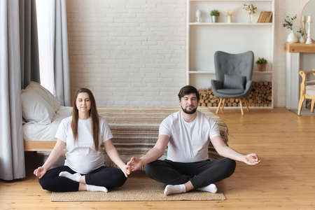 Young pregnant woman practicing yoga with her husband. They meditating in lotus position, their eyes closed. Pregnancy and being maternity and parenthood, wellbeing in family concept. Copy space.の写真素材
