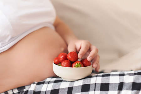Pregnancy, healthy food concept. Beautiful tummy of a pregnant woman, lying on a bed with a strawberries plate. Close up, copy space. Berrys, alergia, diet and healthy food concept, motherhoodの写真素材