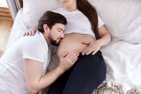 Handsome husband is listening to his beautiful pregnant wife's tummy and smiling, lying together on...