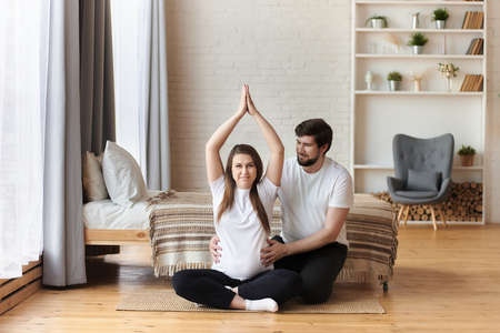 Happy portrait of smiling pregnant woman sitting in lotus position in a bedroom, while her husband gently hugs and supports her. Maternity and parenthood, wellbeing in family concept. Copy space.の写真素材