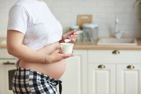 Close-up healthy unknown young pregnant woman in home outfit: a white T-shirt and checkered pants, with cute tummy, holding white greek yogurt for breakfast. Wellbeing at home and pregnancy concept.の写真素材
