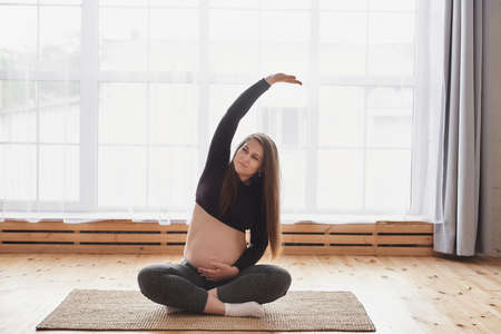 Portrait of smiling young pregnant woman in sports outfit sitting with crossed legs, she holding her tummy and bending aside with raised arm while practicing yoga at home. Motherhood, pregnancy, yogaの写真素材