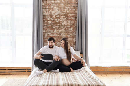 Young man and his pregnant wife using tablet computer on sofa at home. Being maternity and parenthood, wellbeing in family concept. Copy space, internet, modern technology, wireless, be togetherの写真素材
