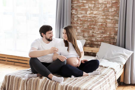 Happy young pregnant woman with her husband holding ultrasound image sitting on a bed at home. Being maternity and parenthood, wellbeing in family concept, pregnancy notice, special family momentsの写真素材