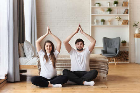Young couple of beautiful pregnant woman and handsome husband practicing yoga and exercise, sitting with hands in Namaste and meditating with closed eyes at home, pregnancy yoga, fitness conceptの写真素材