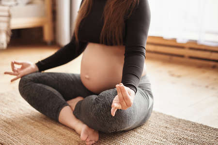 Pregnancy Yoga, Fitness concept. Close-up of beautiful young pregnant yoga model working out indoors. Pregnant woman sitting in yogi cross-legged pose at home. Prenatal meditation, Motherhoodの写真素材