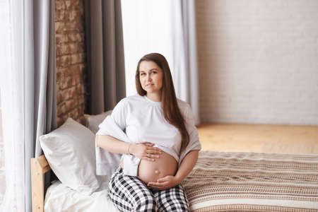 Calm pregnant woman looking at camera in home outfit: a white T-shirt and checkered pants sits on the bed, hugs her tummy, dreams of future motherhood. Prenatal, Baby expecting concept. Copy space.の写真素材