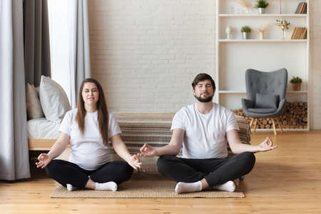 Young couple of beautiful pregnant woman practicing yoga, meditating in lotus position with closed eyes in a bedroom. Pregnancy and being maternity and parenthood, wellbeing in family concept, fitnessの写真素材