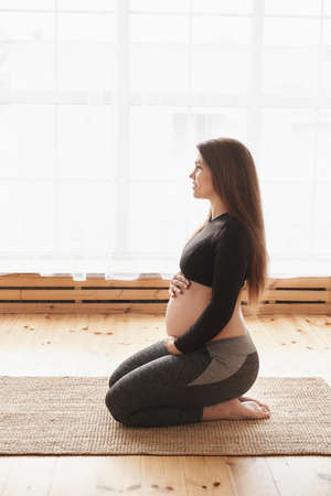 Side view of young pregnant woman sits on her knees on mat doing hip stretching exercises, hugs her tummy, looks in front of her. Vertical, Full length, copy space, Pregnancy Yoga and Fitness conceptの写真素材