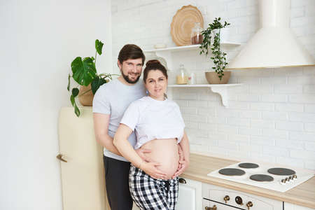 Future parents in home outfit embrace standing in the kitchen, looking at camera. Healthy Lifestyle,...