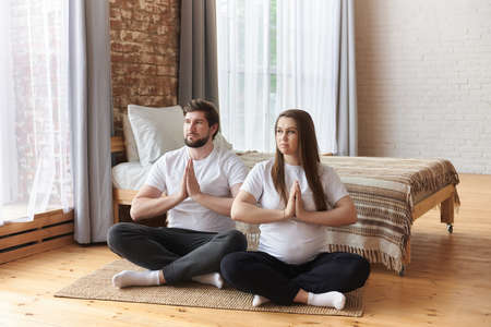 Young couple of beautiful pregnant woman and handsome husband practicing yoga and exercise, sitting with hands in Namaste, and meditating at home. Prenatal Yoga. Healthy maternity concept, fitnessの写真素材