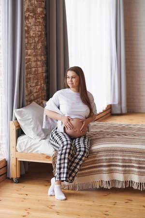 Happy smiling pregnant woman looking away in home outfit: a white T-shirt and checkered pants sits on the bed, hugs her tummy, dreams of future motherhood. Prenatal, Baby expecting concept, full lengthの写真素材