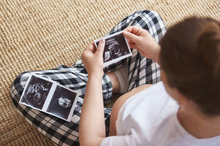 Young mother waiting of the baby. Pregnant woman sitting on a rug, holding ultrasound image. Concept of pregnancy, health care, gynecology, medicine. Close-up, copy space, indoors, top viewの写真素材