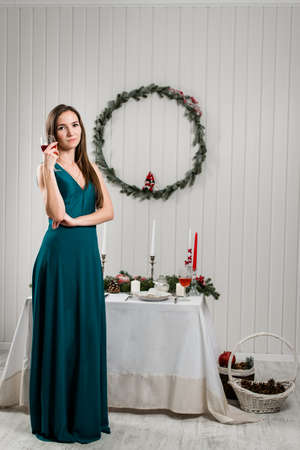 Winter holidays celebration. A picture of a tall girl full height in a long dress with a wine glass opposite a Christmas decorated table. Woman Enjoying Christmas Dinner. He looks at the camera.の写真素材